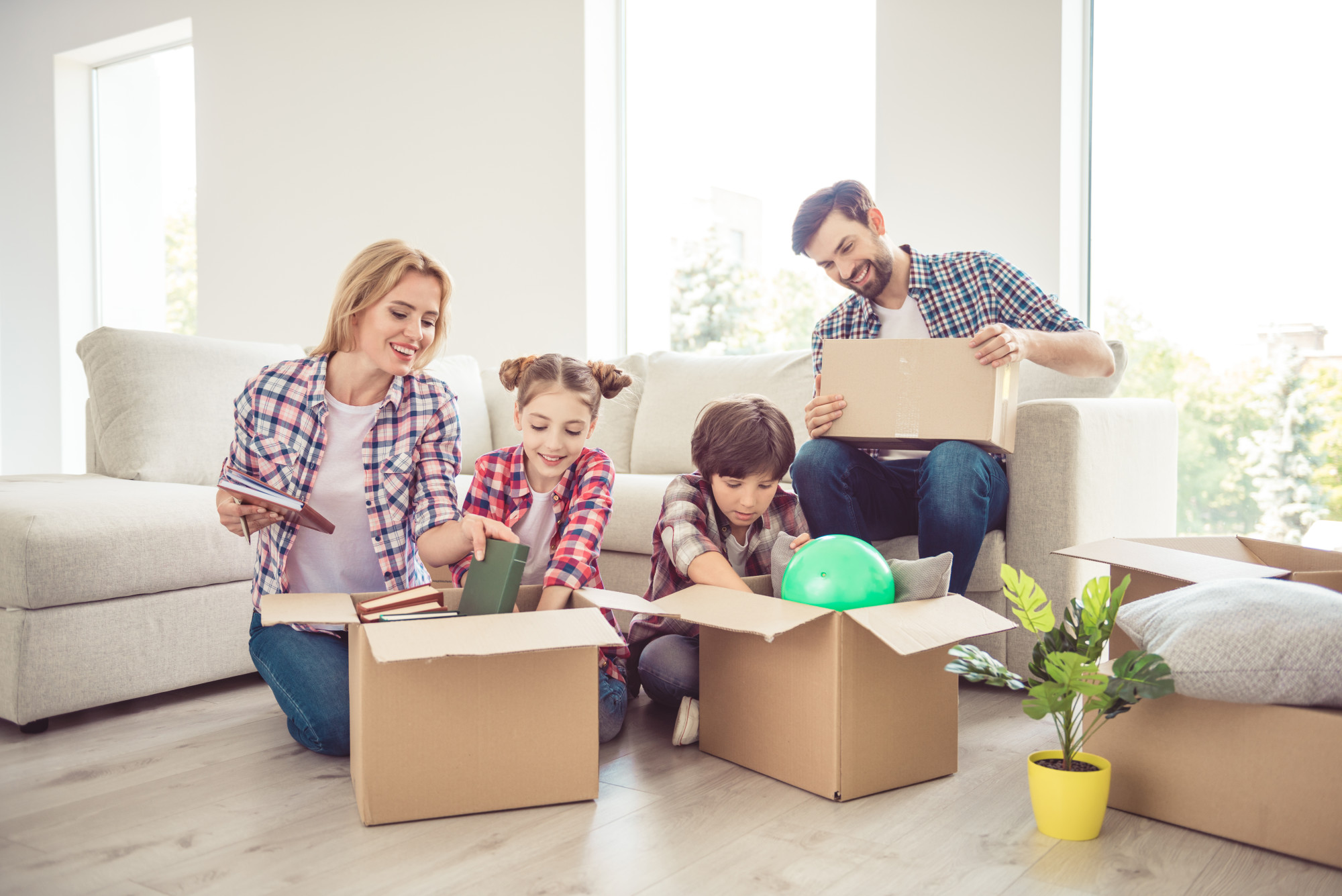 Young happy smiling family four persons unwrapping carton boxes with stuff in light living room, moving to new flat, renovation