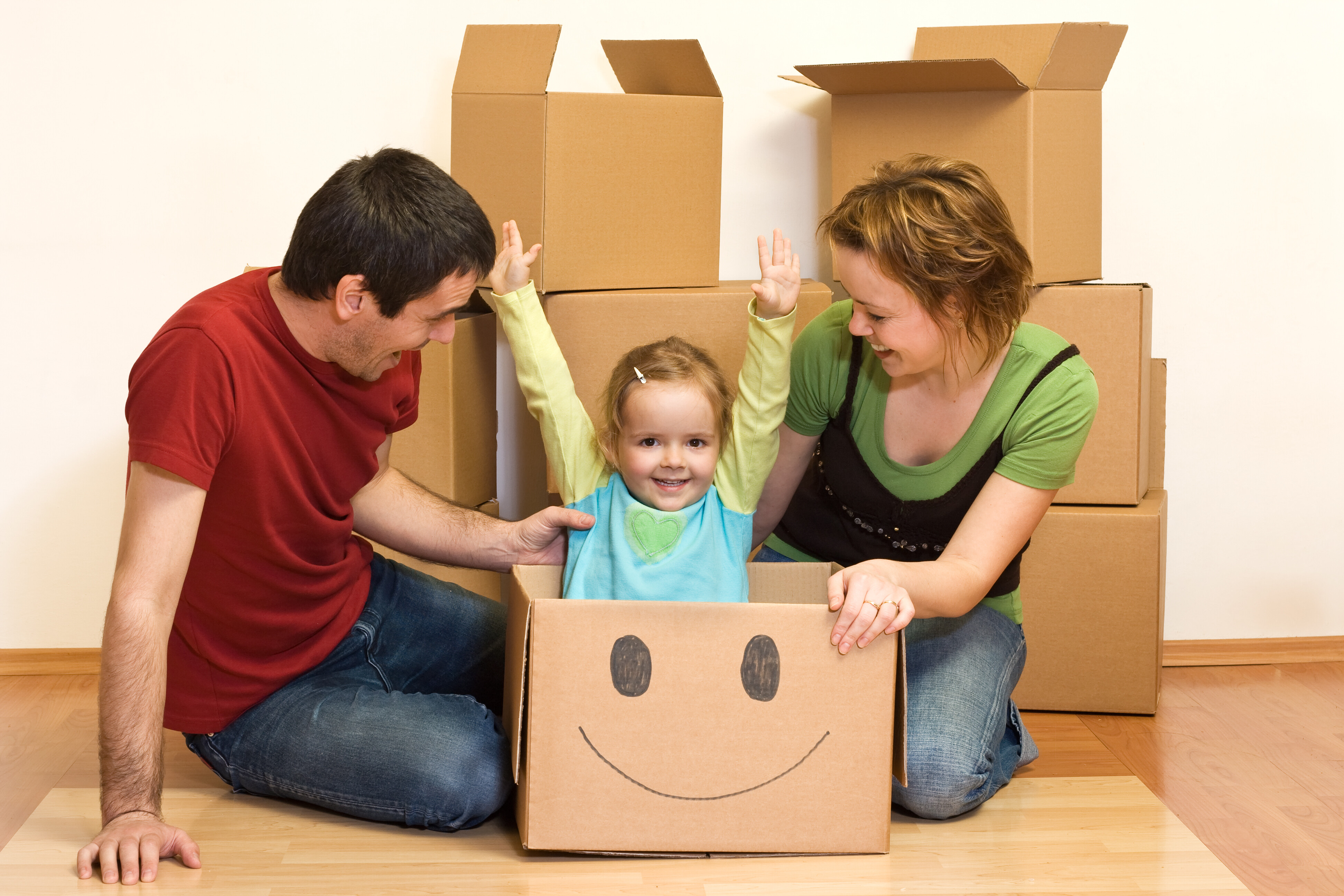 Woman and man with happy small child sitting in a moving box. Box has a smiley face on it and child has arms raised in the air.