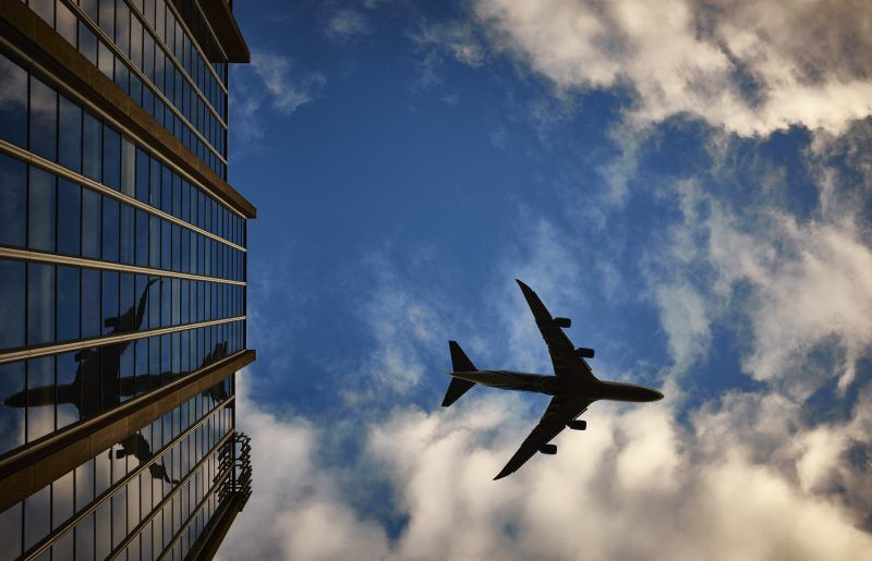 Airplane flying above reflecting in glass building against cloudy sky.